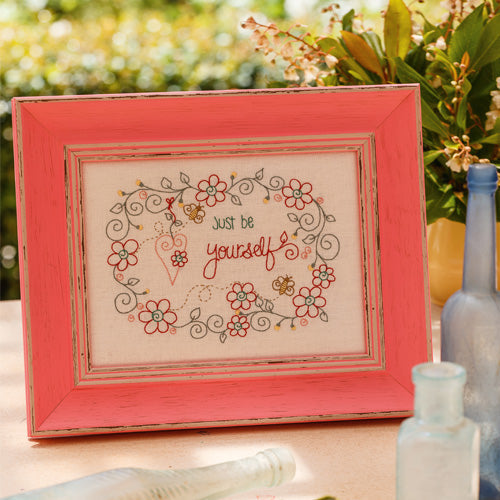 Styled shot of framed embroidery with flowers, hearts and bees around words 'just be yourself'